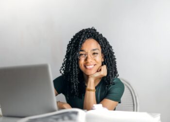 Joyful businesswoman with curly hair smiling at camera while using laptop indoors.
