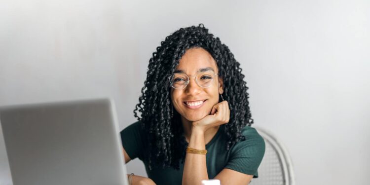 Joyful businesswoman with curly hair smiling at camera while using laptop indoors.