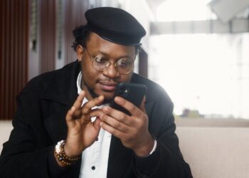 A cheerful man wearing a stylish outfit uses a smartphone while seated in a modern indoor setting.