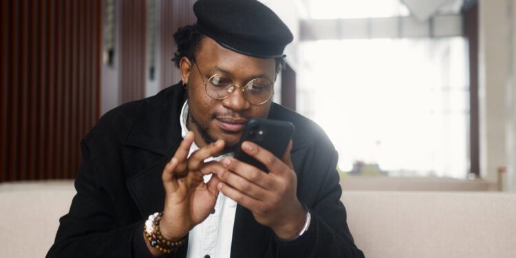 A cheerful man wearing a stylish outfit uses a smartphone while seated in a modern indoor setting.